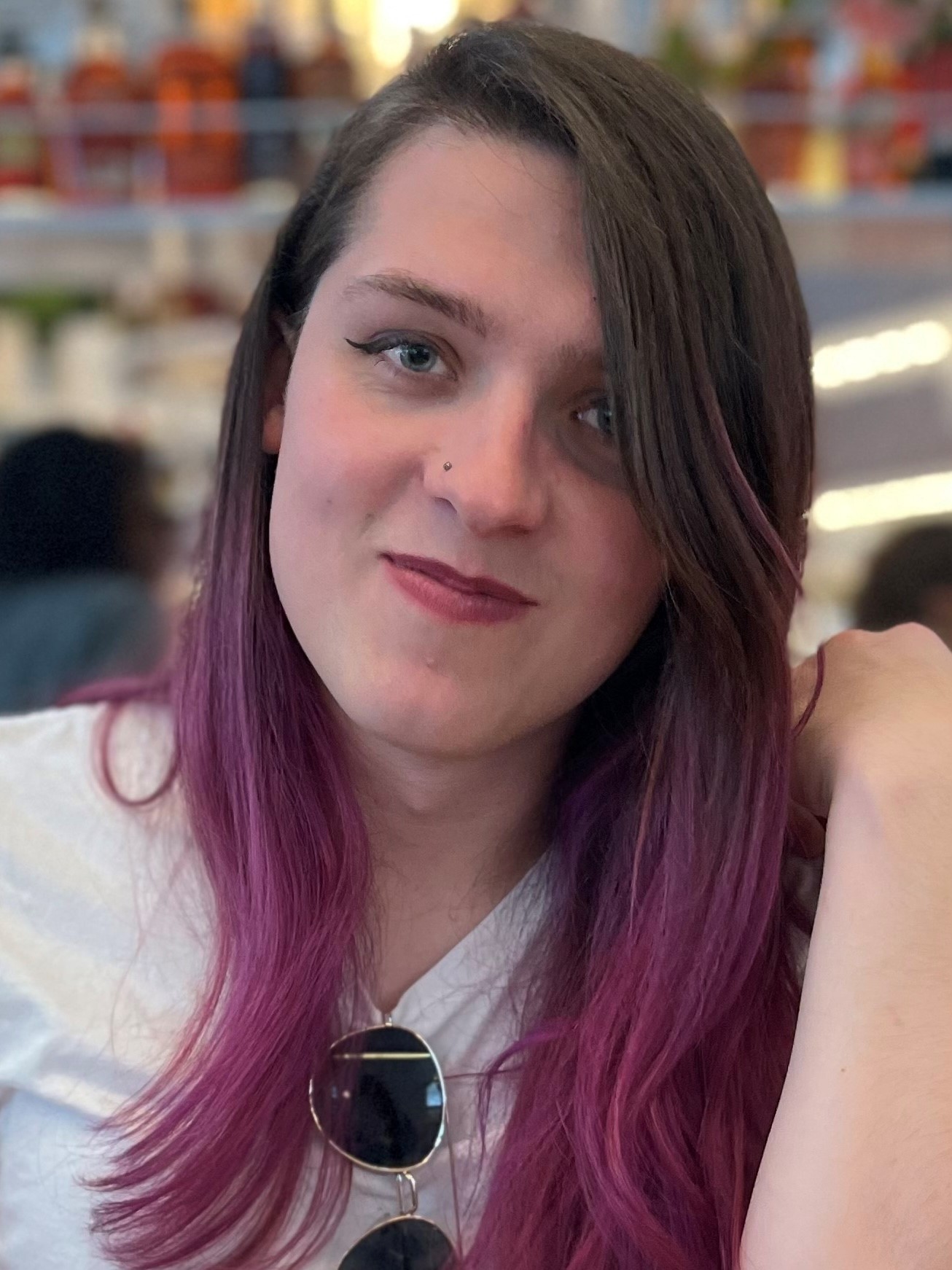 Person with long brown and pink hair smiles into the camera. She has sunglasses hanging on the front of her shirt. 