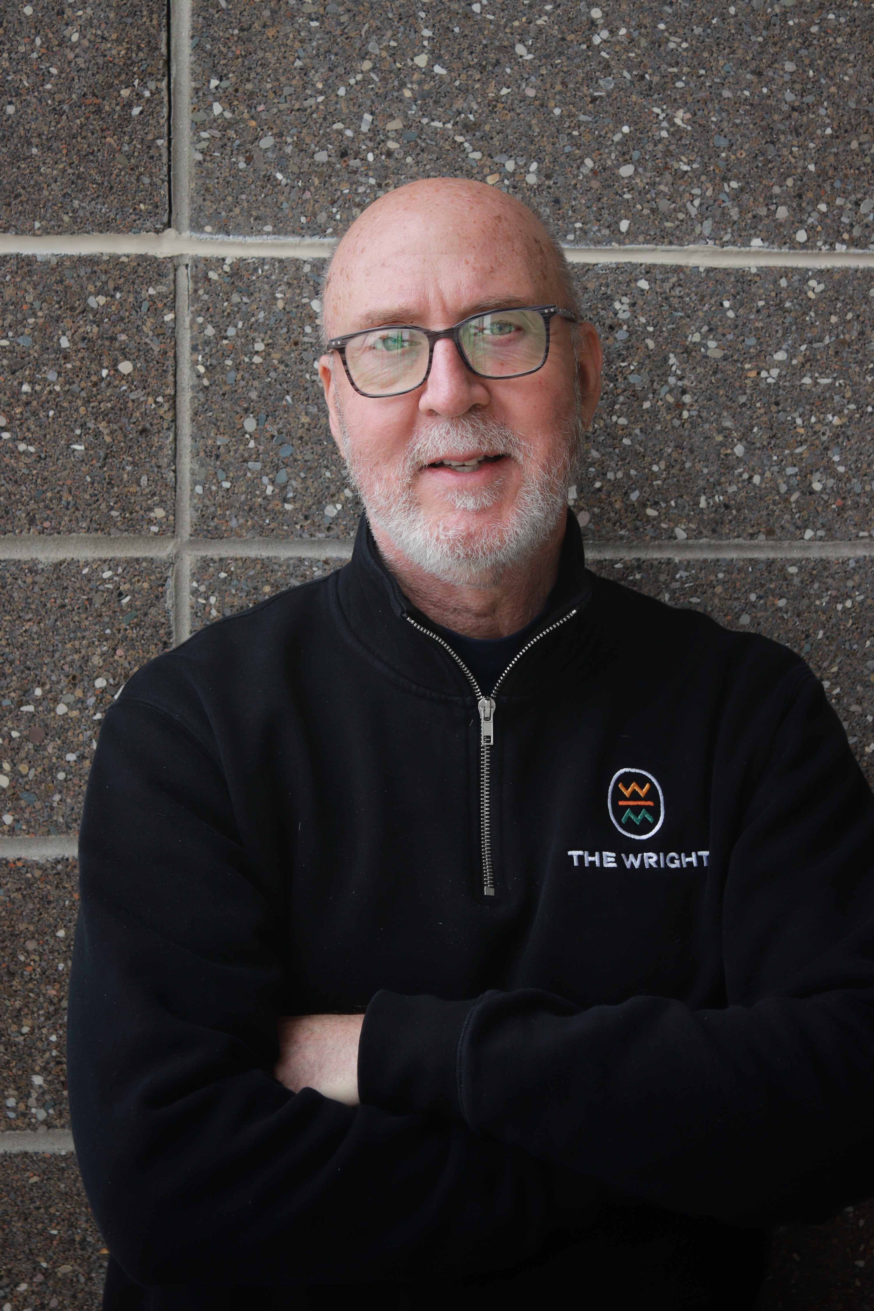 Man with glasses looks at the camera. He is standing in front of a gray brick wall and wearing a fleece that reads "the wright." 