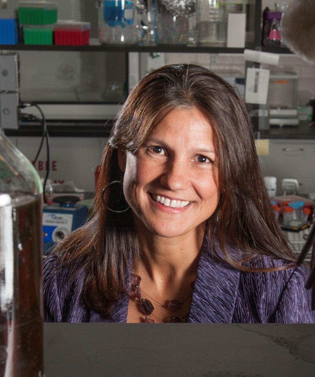 Woman with long brown hair and silver hoop earrings smiles at the camera. She is in a lab.